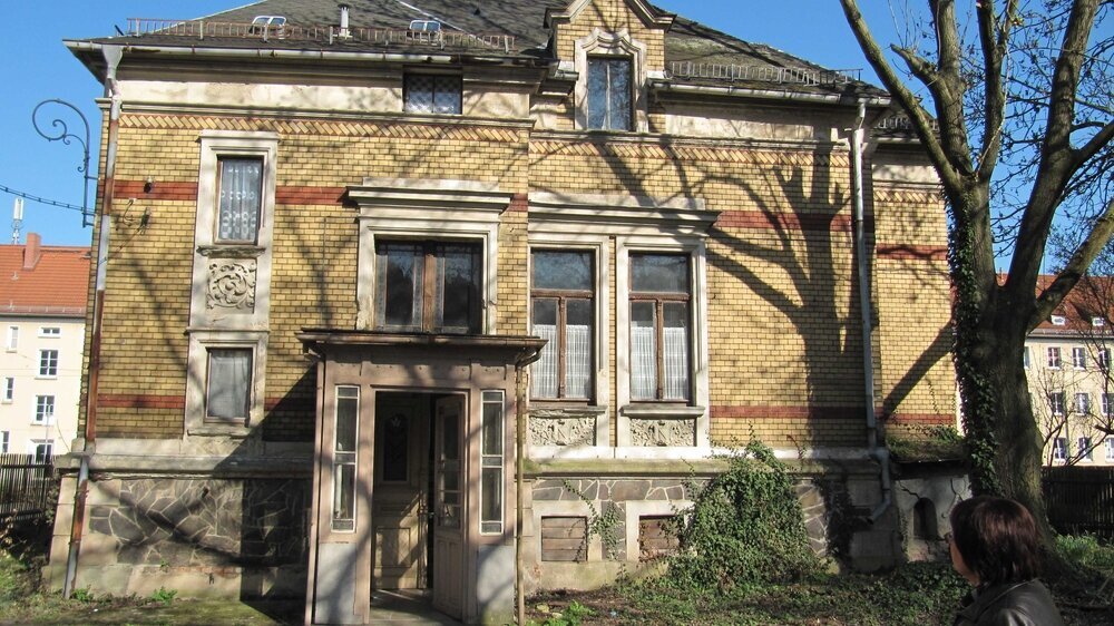 Die Villa, wie sie Schneeweiß gekauft hat, von der Hofseite aus gesehen. "Der Parozustand des Hauses, also quasi der Halteapparat, sprich der Keller, bestand nur aus lose gesetztem Bruchsteinwerk, die Stahlträger der Kappendecken waren an den Übergäng Die Villa, wie sie Schneeweiß gekauft hat, von der Hofseite aus gesehen. "Der Parozustand des Hauses, also quasi der Halteapparat, sprich der Keller, bestand nur aus lose gesetztem Bruchsteinwerk, die Stahlträger der Kappendecken waren an den Übergängen zur Außenwand durch den Rost wie Blätterteig aufgeblättert. Eine Überraschung folgte auf die nächste, aber es gab ja kein Zurück mehr.\r\n"Da lacht das Herz eines Restaurators"\r\n Für mich war die Abgeschlossenheit des alten Herrn jedoch ein Glücksumstand, kein Handwerker hat ihm Pressspantüren mit Plasteklinken, Plastefenster und abgehängte Gipskartondecken aufgeschwatzt. Unter mehreren Farbschichten kamen Türen mit schöner Bierlasur, die originalen Messingklinken und -griffe zum Vorschein. Da lacht doch das Herz eines Restaurators und Denkmalpflegers, sowie das Herz des Zahnarztes lacht, wenn sich ein scheinbar sklerosierter und obendrein stark gekrümmter Wurzelkanal doch bis zur Iso-Größe 30 punktgenau bis zum Apex aufbereiten lässt ... Es schloss sich eine aufwendige Handwerkersuche an, der Nachwuchsmangel betrifft ja nicht nur die Zahnärzte, sondern auch viele Handwerksbetriebe. Der schieferverarbeitende Dachdecker kam aus Sachsen, der Schmied aus Sachsen-Anhalt, der Stuckateur aus dem fernen Apolda. Die Sanierung der alten Villa in Anlehnung an die denkmalpflegerische Zielsetzung dauerte letztendlich etwa fünf Jahre, drei DIN-A4-Seiten mit Auflagen wollten eingehalten werden und die parallel begonnene Promotion ganz nebenbei auch noch abgeschlossen werden."