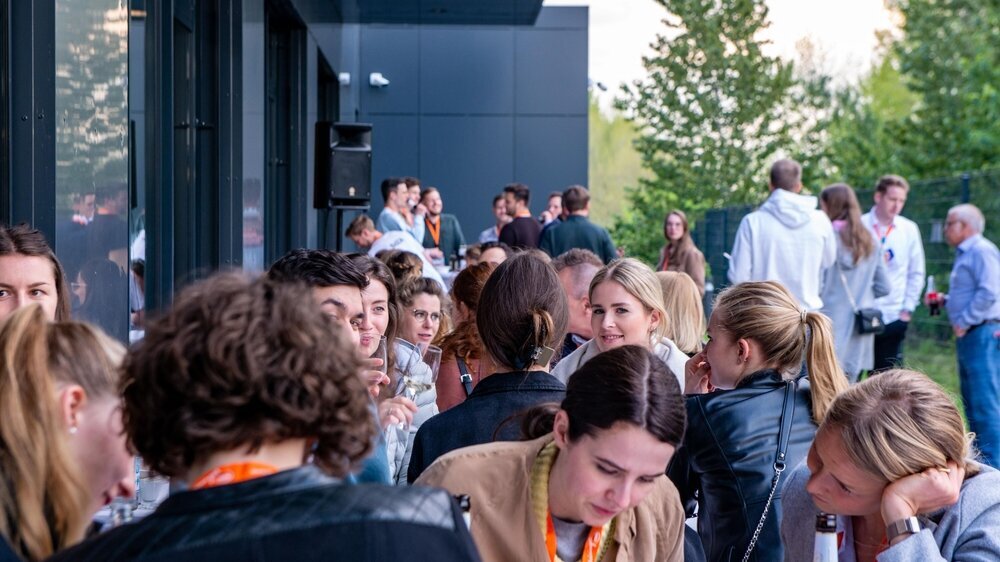 Get together bei Getränken auf der AluFaTa 2023 Eine Gruppe von circa 70 jungen Erwachsenen sitzen im Rahmen der Alumnifachtagung auf einer Terrasse zusammen und unterhalten sich.