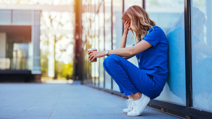 Tired nurse sits on floor outdoors of the medicine clinic after a hard dutty. Exhausted tired doctor or nurse. Clinic and hospital medical stuff working over hours. Overworked professional. Erschöpfte Krankenhausmitarbeiterin in der Kaffeepause