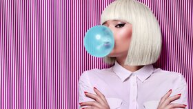 Junge Frau mit weißer Perücke bläst blaue Kaugummiblase vor gestreiftem Hintergrund.