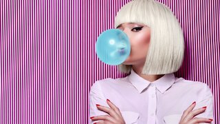 Junge Frau mit weißer Perücke bläst blaue Kaugummiblase vor gestreiftem Hintergrund.