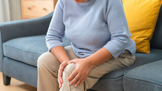 Body arthritis muscles stiff problem, aged woman suffering on leg, pain knee from disease chronic, hand massage rubbing joint injury, mature hurt or sore sitting on sofa at home. Health care people.