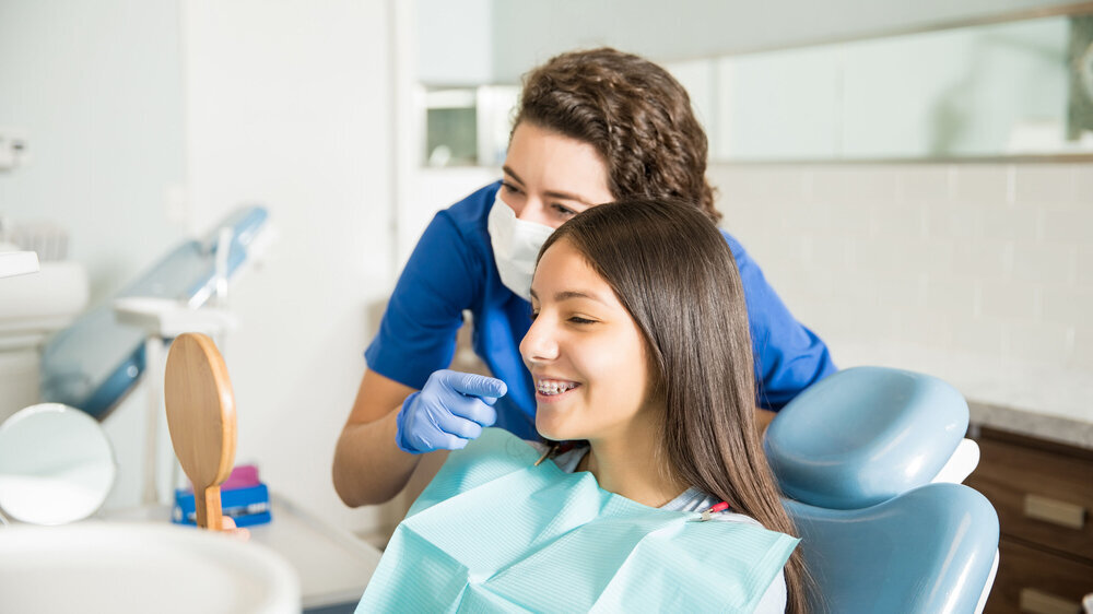 Female dentist showing braces to teenage girl in mirror at dental clinic