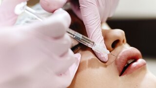The process of lips enhancement. Cosmetician makes an injection of hyaluronic acid in a beautiful lips. The young girl with a beautiful face in the special hat and hands of the doctor in the pink gloves.