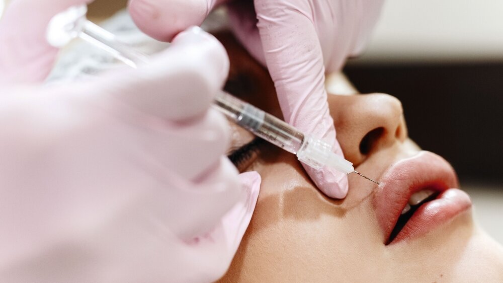 The process of lips enhancement. Cosmetician makes an injection of hyaluronic acid in a beautiful lips. The young girl with a beautiful face in the special hat and hands of the doctor in the pink glov The process of lips enhancement. Cosmetician makes an injection of hyaluronic acid in a beautiful lips. The young girl with a beautiful face in the special hat and hands of the doctor in the pink gloves.