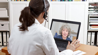 Female doctor of geriatrics in her surgery office with headset in front of her laptop talking via video call with an old patient about her prescribed drugs