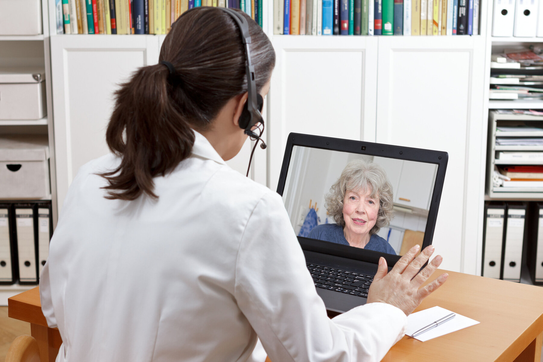 584325-flexible-1900.jpg Female doctor of geriatrics in her surgery office with headset in front of her laptop talking via video call with an old patient about her prescribed drugs