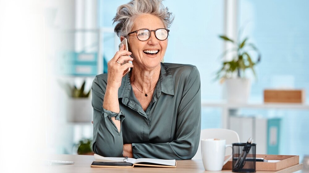 Ältere Angestellte lacht mit Handy in der Hand, freundliche Ausstrahlung am Arbeitsplatz. Ältere Angestellte lacht mit Handy in der Hand, freundliche Ausstrahlung am Arbeitsplatz.