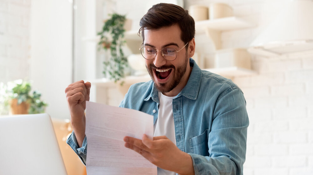 Excited man reading postal mail letter overjoyed by good news Excited euphoric happy young man holding reading paper postal mail letter amazed overjoyed by good news, got new job celebrate taxes refund receive salary payment loan approval sit at home table