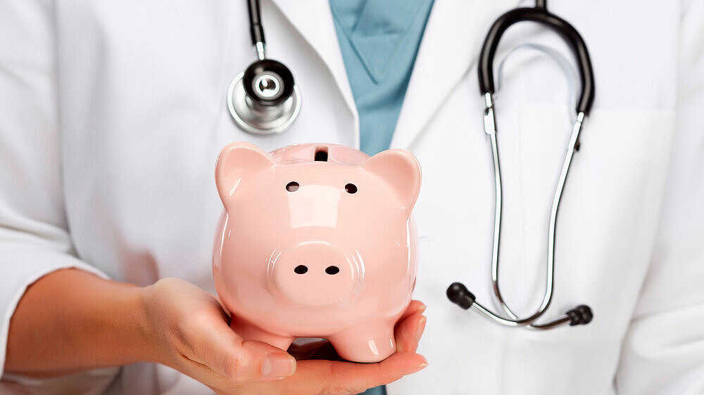 Arzt hält Sparschwein in der Hand als Symbol für die Kosten im Gesundheitswesen.