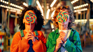 Zwei Kinder stehen auf einem Jahrmarkt und halten knallbunte Zuckerlollis vor ihre Gesichter.