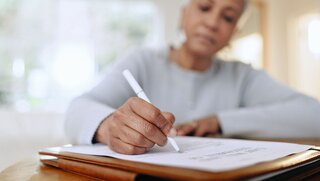 Senior woman, sign and writing with documents, paperwork and application for life insurance policy. Person, hand and checklist for compliance, investment or will in retirement with signature in home.