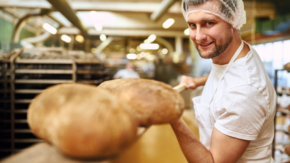 Bäcker Lehrling mit fertig gebackenem Brot auf Brotschaufel Junger Mann als Bäcker Lehrling mit fertig gebackenem Brot auf Brotschaufel in der Bäckerei