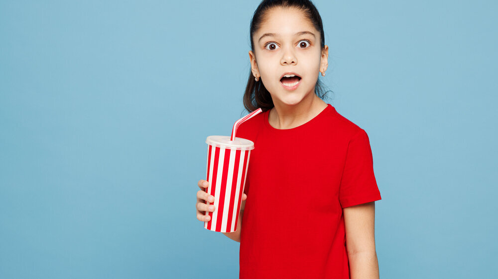 Little shocked child kid girl 8-9 years old wear red t-shirt have fun hold in hand cup of soda pop cola fizzy water isolated on blue color background studio Mother's Day love family lifestyle concept Little shocked child kid girl 8-9 years old wears red t-shirt have fun hold in hand cup of soda pop cola fizzy water isolated on blue color background studio Mother's Day love family lifestyle concept