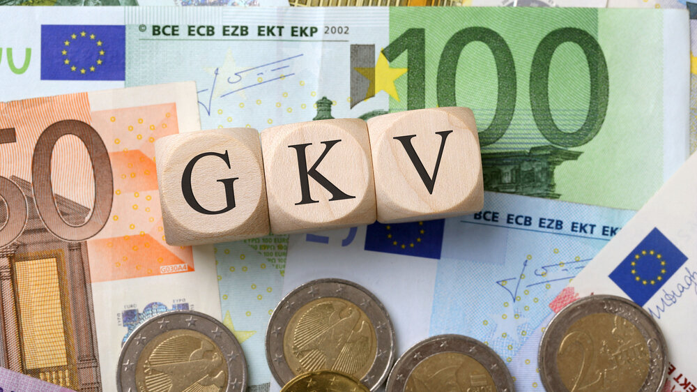 Auf einer Ansammlung von Eurobanknoten und Euromünzen liegen drei hellbraune Holzwürfel, die zusammen das Wort GKV ergeben.
