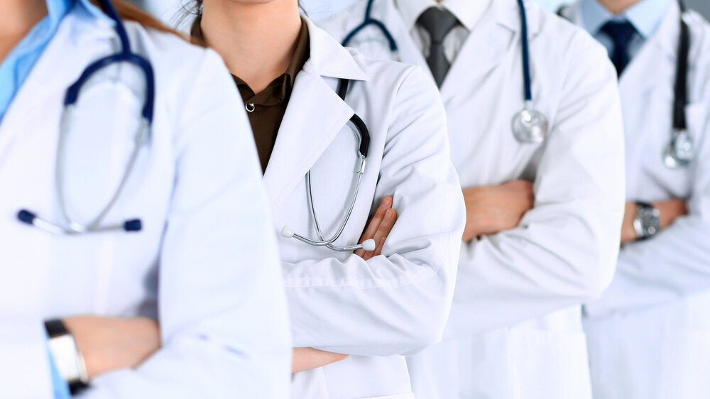 Eine Gruppe Ärztinnen und Ärzte steht als Team mit verschränkten Armen in einem Krankenhausbüro. Eine Gruppe Ärztinnen und Ärzte steht als Team mit verschränkten Armen in einem Krankenhausbüro.