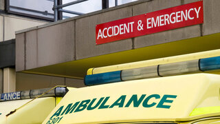 The cabs of two yellow ambulances parked below an Accident & Emergency department sign outside an NHS hospital in the UK