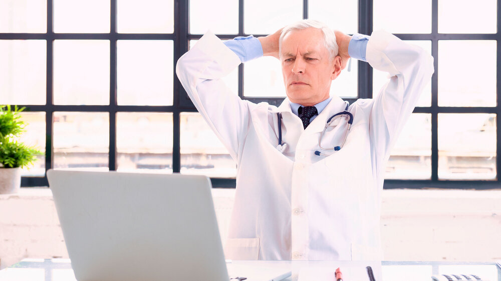 Thinking male doctor sitting in doctor's office behind his laptop Shot of careworn male doctor sitting behind his laptop in doctor’s office.
