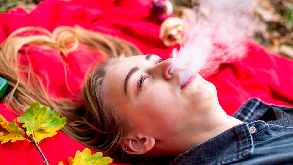 Vape teenager. Young beautiful white teenage girl in casual clothes lies on a red plaid and vapes electronic cigarette on the street in the park in the autumn evening. Bad habit. Vaping dampfender Teenager