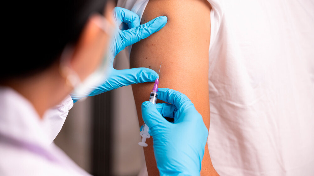 Arzthand in blauem Handschuhen hält Spitze mit Impfstoff zur Injektion in die Schulter eines Patienten.