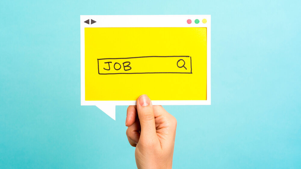 Symbolbild für die Jobsuche im Internet Vor einem hellblauen Hintergrund hält eine Hand ein gelbes Papier mit einer gezeichneten Suchmaske hoch, in der "Job" geschrieben steht.