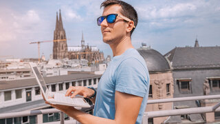 A young man remotely works on a laptop on an observation deck against the background of a view of Cologne Cathedral in Germany. Freelancer and student online concept