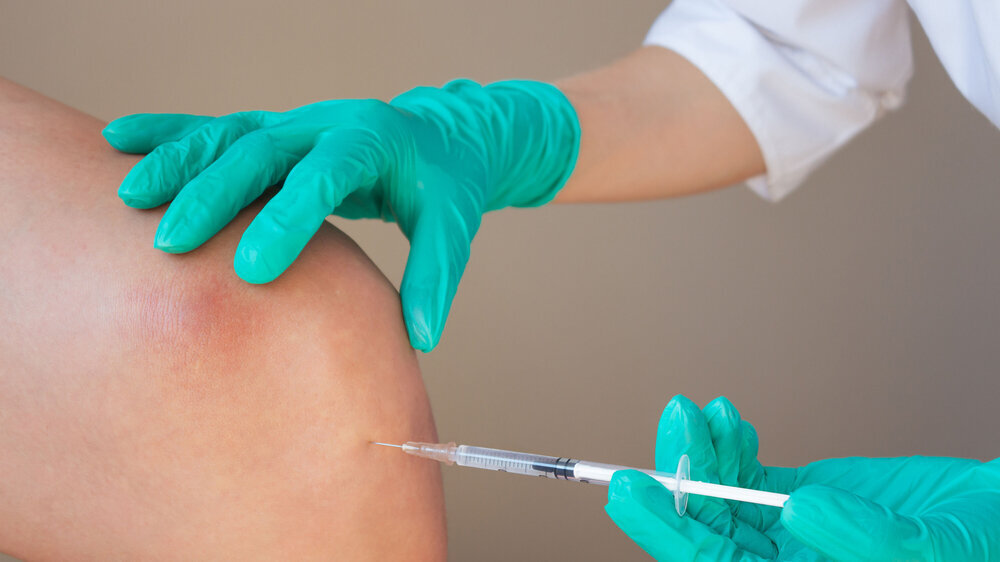 A doctor in green gloves gives a young man an intra-articular injection of hyaluronic acid against osteoarthritis in the knee joint. Preventing the destruction of the knee joint. A doctor in green gloves gives a young man an intra-articular injection of hyaluronic acid against osteoarthritis in the knee joint. Preventing the destruction of the knee joint.