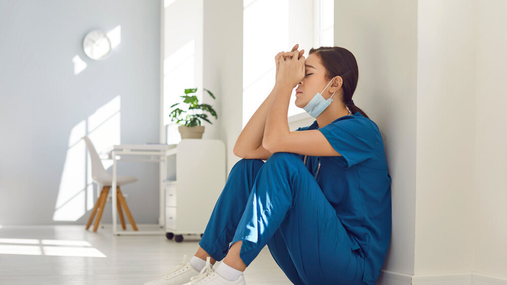 Müde, deprimierte Krankenschwester mit blauer Gesichtsmaske und Uniform sitzt auf dem Krankenhausboden. Müde, deprimierte Krankenschwester mit blauer Gesichtsmaske und Uniform sitzt auf dem Krankenhausboden.