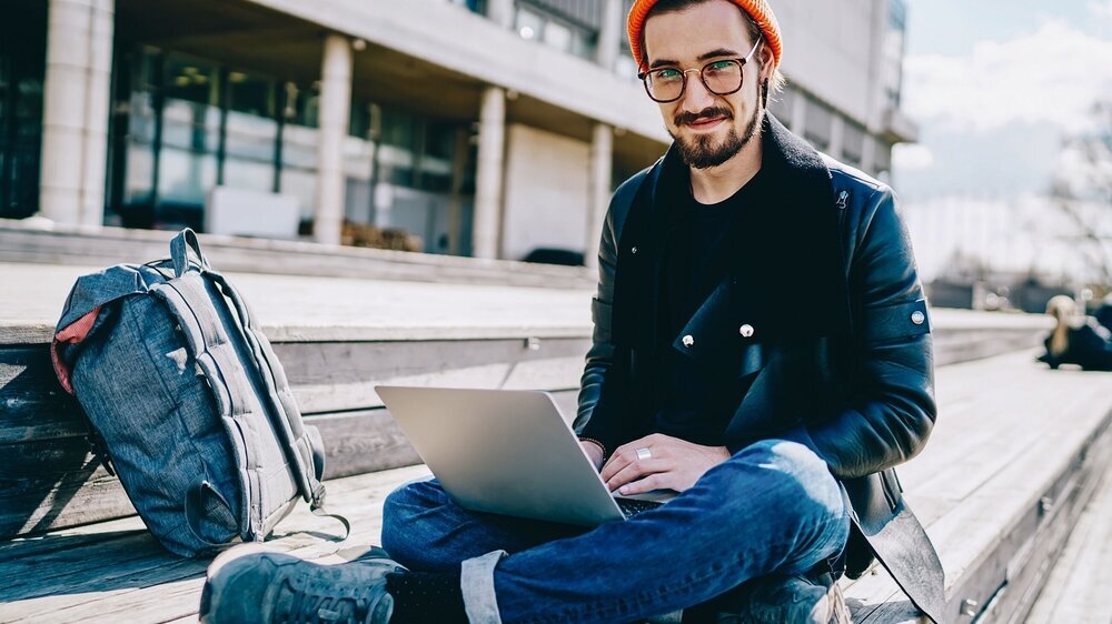 Portrait of smiling trendy dressed male student in spectacles using laptop computer for networking and learning, hipster guy looking at camera sitting with netbook connected to 4G internet on street. Generation Z Mann mit Laptop