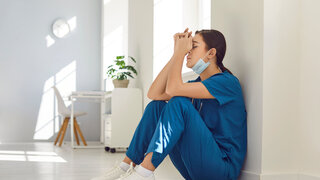 Müde, deprimierte Krankenschwester mit blauer Gesichtsmaske und Uniform sitzt auf dem Krankenhausboden.