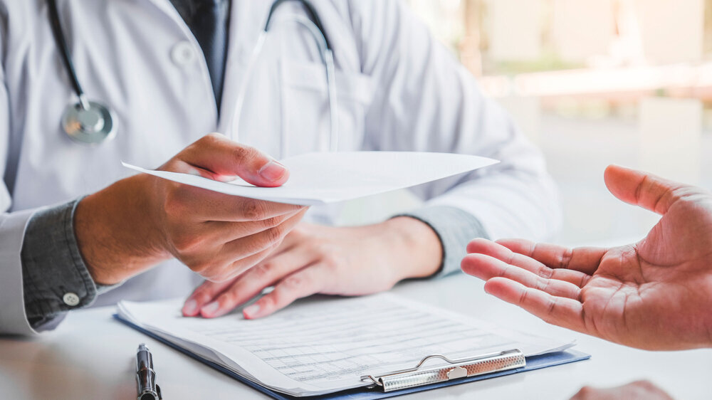 Arzt stellt Patienten Rezept aus und reicht es ihm über den Schreibtisch in der Praxis.