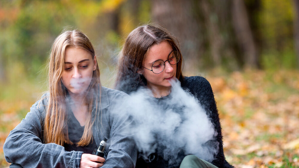 Vape lgbt teenagers. Bisexual lesbian young caucasian teenage girls in casual clothes vape electronic cigarette on the street in the park in the autumn evening. Bad habit. Vape lgbt teenagers. Bisexual lesbian young caucasian teenage girls in casual clothes vape electronic cigarette on the street in the park in the autumn evening. Bad habit.