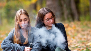 Vape lgbt teenagers. Bisexual lesbian young caucasian teenage girls in casual clothes vape electronic cigarette on the street in the park in the autumn evening. Bad habit.