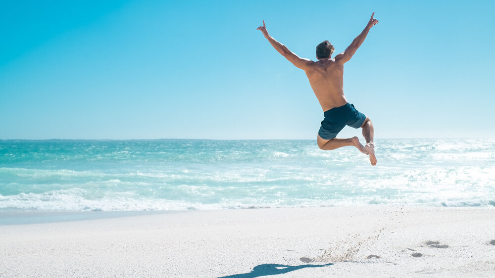 Rückansicht eines jungen Mannes, der mit ausgestreckten Armen am Strand freudig in die Luft springt. Rückansicht eines jungen Mannes, der mit ausgestreckten Armen am Strand freudig in die Luft springt.