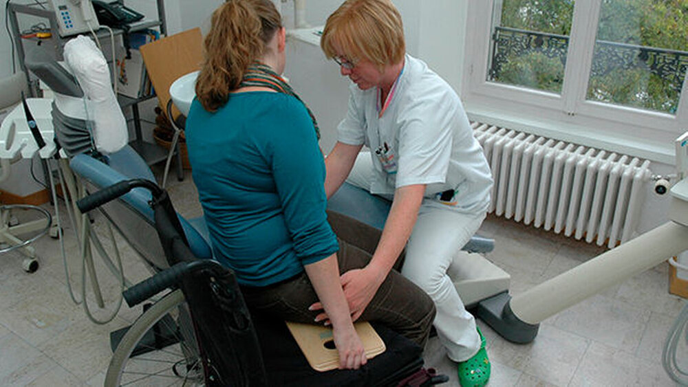 Abb. 9: Das Brett wird etwas unter das Gesäß der Patientin geschoben. Die betreuende Person setzt sich auf das Fußende des Behandlungsstuhls und fasst die Patientin an der Hüfte. Nun kann die Patientin – mit eigener Unterstützung – langsam auf den Behandlungsstuhl bewegt werden. Die Patientin hält dabei das Brett am Griff fest. |