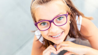 Young preteen girl in glasses wearing braces smiles at the camera showing heart shape with her hands.