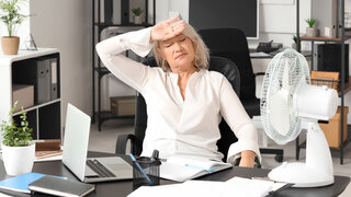 Mature businesswoman suffering from hot flashes at table in office. Menopause concept