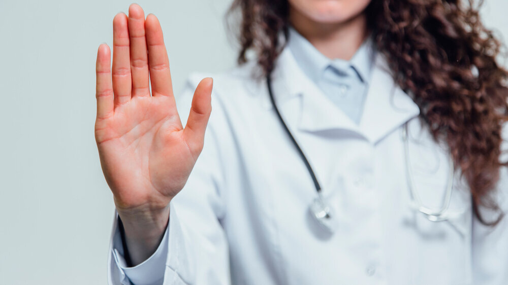 Junge Ärztin hält schützend die Hand erhoben, um Gewalt seitens Patienten abzuwehren.