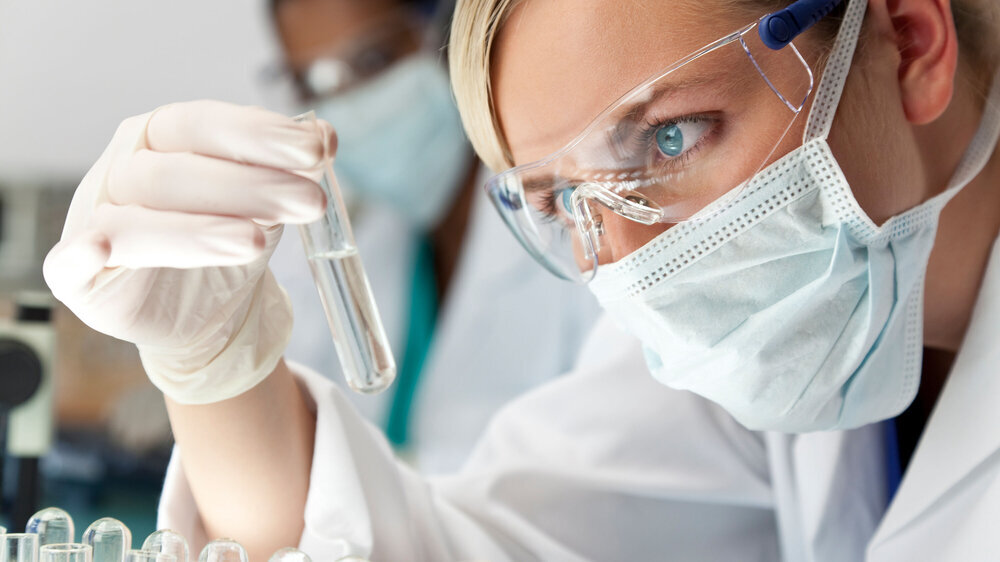 118297969 A blond medical or scientific researcher or doctor using looking at a clear solution in a laboratory with her Asian female colleague out of focus behind her.