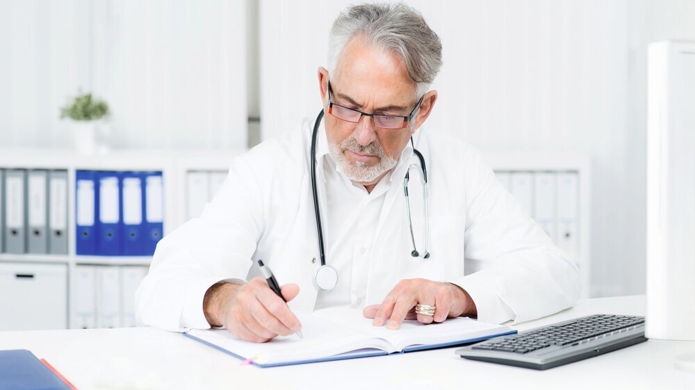 Älterer Hausarzt am Schreibtisch seiner Praxis.