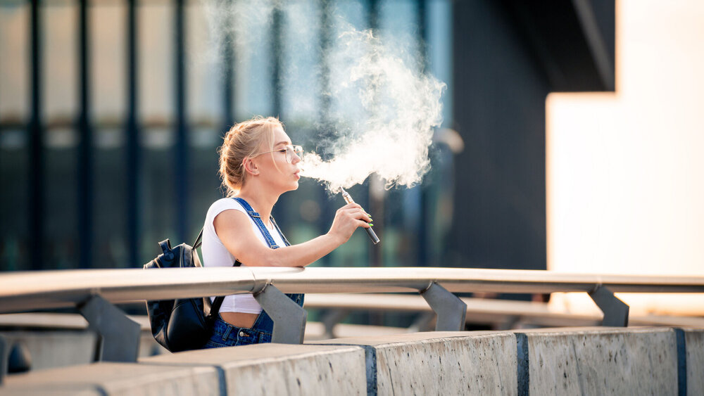 Young woman outdoor vaping e-cigarette on modern city buildings background