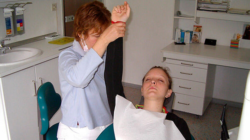 Der Arm der Patientin wird dazu hochgestellt. Das sich infolge der Kältesuggestionen ausbreitende Taubheitsgefühl in der linken Hand wird auf die Behandlungszone im Mundbereich hypnotisch übertragen.