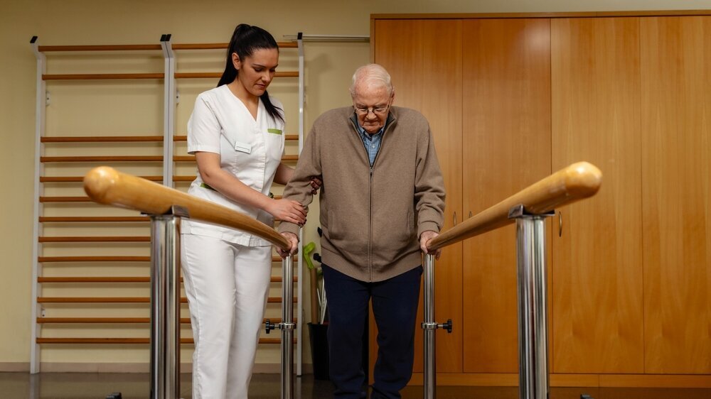 Senior stroke patient doing rehabilitation in clinic with parallel bars. Gull length portrait of young nurse assisting senior stroke patient in clinic. Old man doing rehabilitation with parallel bars.