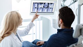 Zahnärztin zeigt im Gespräch mit einem Patienten auf einen Monitor
