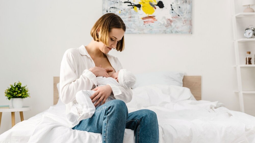 Mother in jeans sitting on bed and breastfeeding baby Stillen in Jeans auf Bett