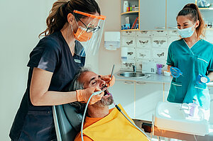 Männlicher Patient, der auf einem Zahnarztstuhl sitzt und von einem Zahnarzt mit Unterstützung einer Dentalhygienikerin eine Zahnsanierung erhält Männlicher Patient, der auf einem Zahnarztstuhl sitzt und von einem Zahnarzt mit Unterstützung einer Dentalhygienikerin eine Zahnsanierung erhält
