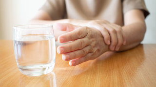 Elderly woman hands tremor symptom reaching out for a glass of water on wood table