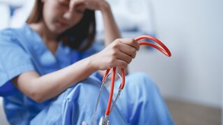 Junge Ärztin in blauem Kittel fühlt sich überwältigt und gestresst, während sie in einem Krankenhauszimmer auf dem Boden sitzt