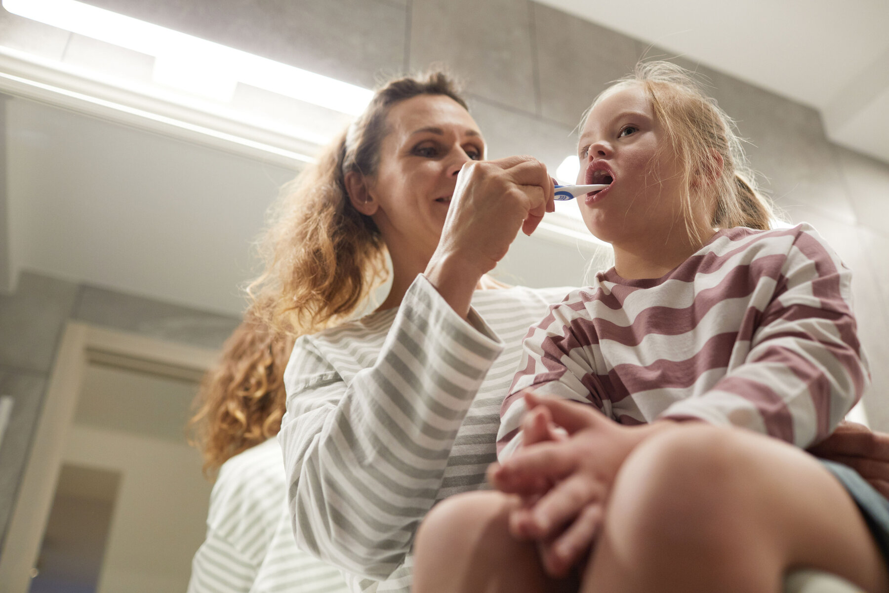 Low angle portrait of smiling mother helping little girl with down syndrome brush teeth in bathroom, copy space