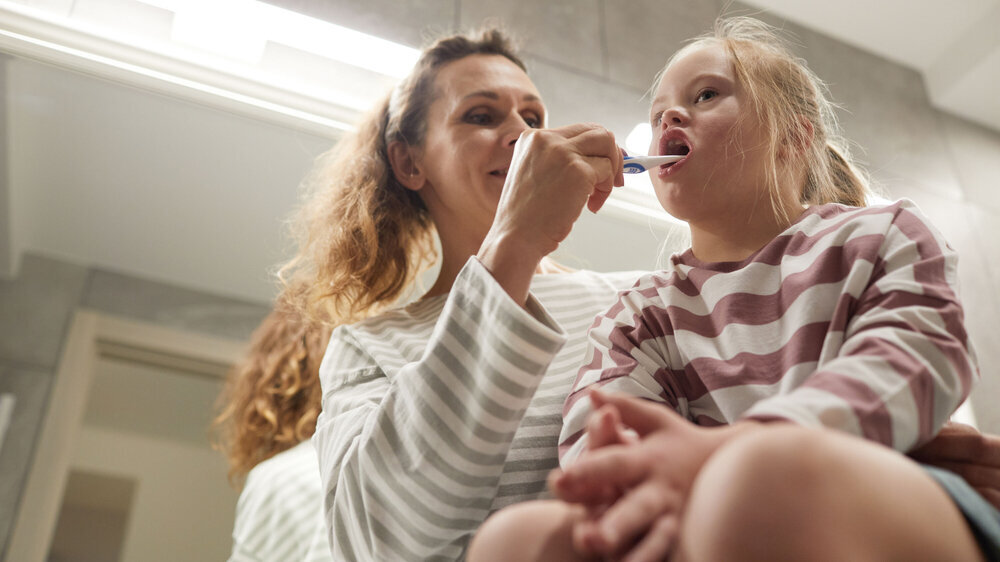 Mother Helping Child with Special Needs Low angle portrait of smiling mother helping little girl with down syndrome brush teeth in bathroom, copy space